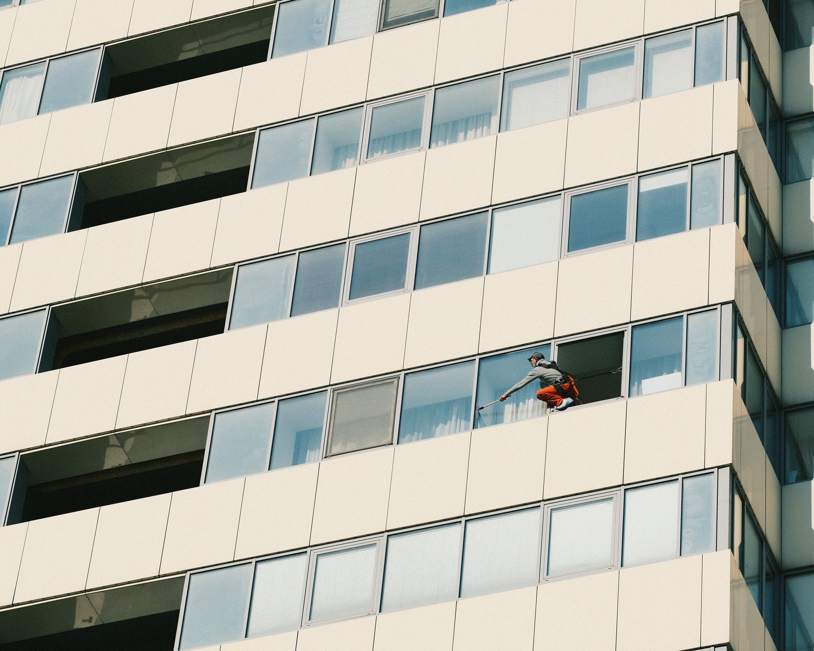 A window cleaner suspended by rope harness cleaning the exterior glass facade of a tall modern high-rise building