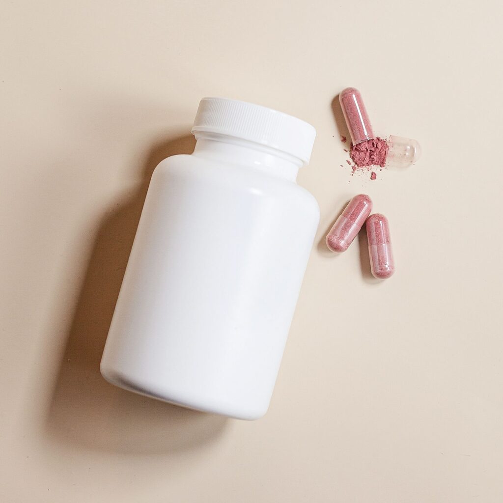 Overhead view of a plain white supplement bottle with pink capsules spilling onto a beige surface