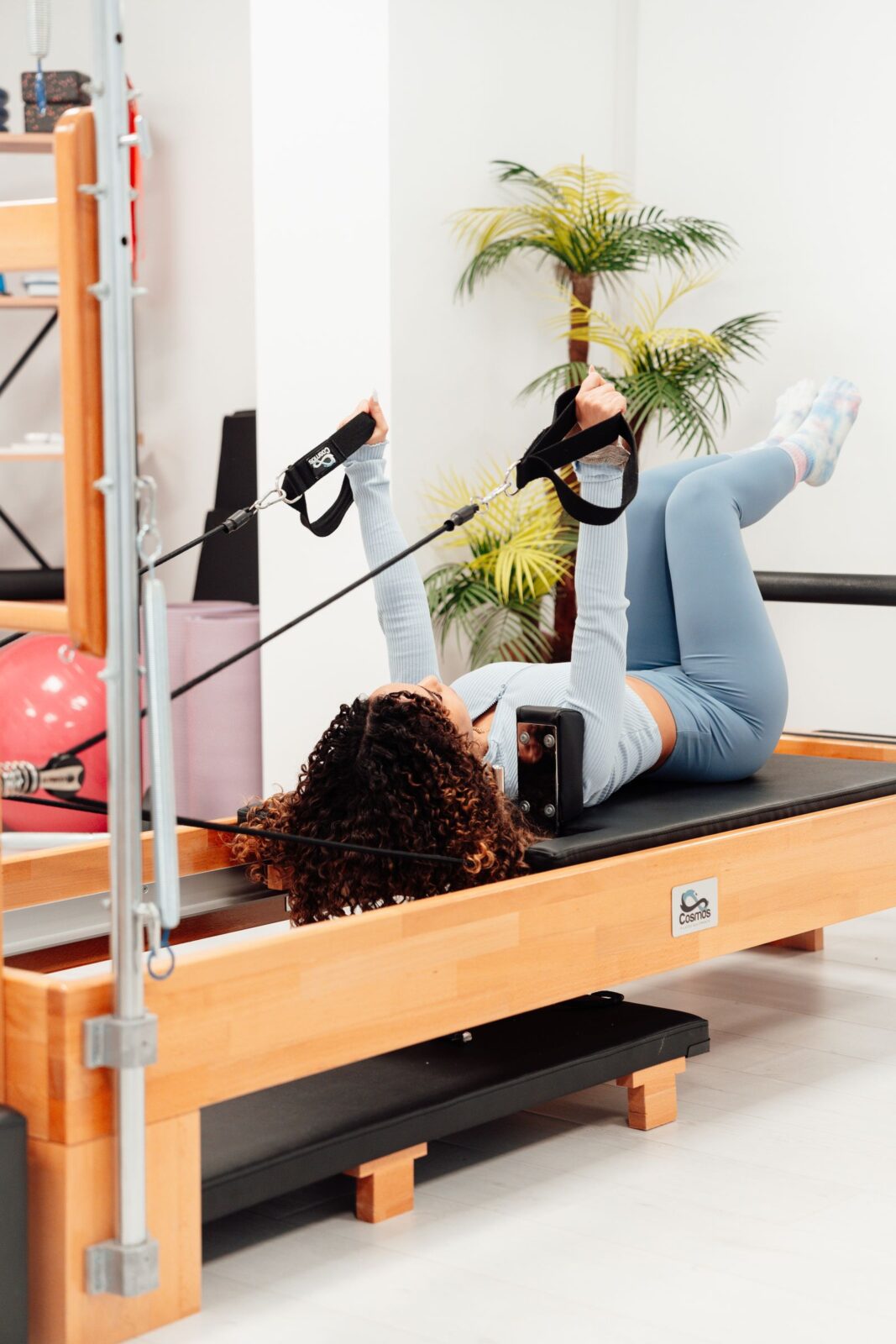 A woman exercising on a Pilates reformer, pulling the carriage using the ropes and straps