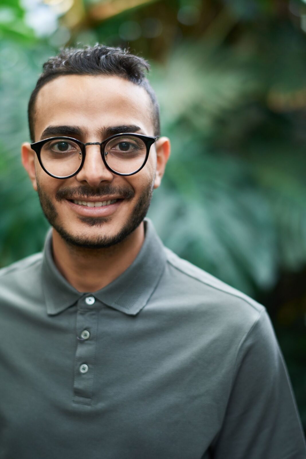 A confident man wearing glasses smiles outdoors with lush foliage in the background