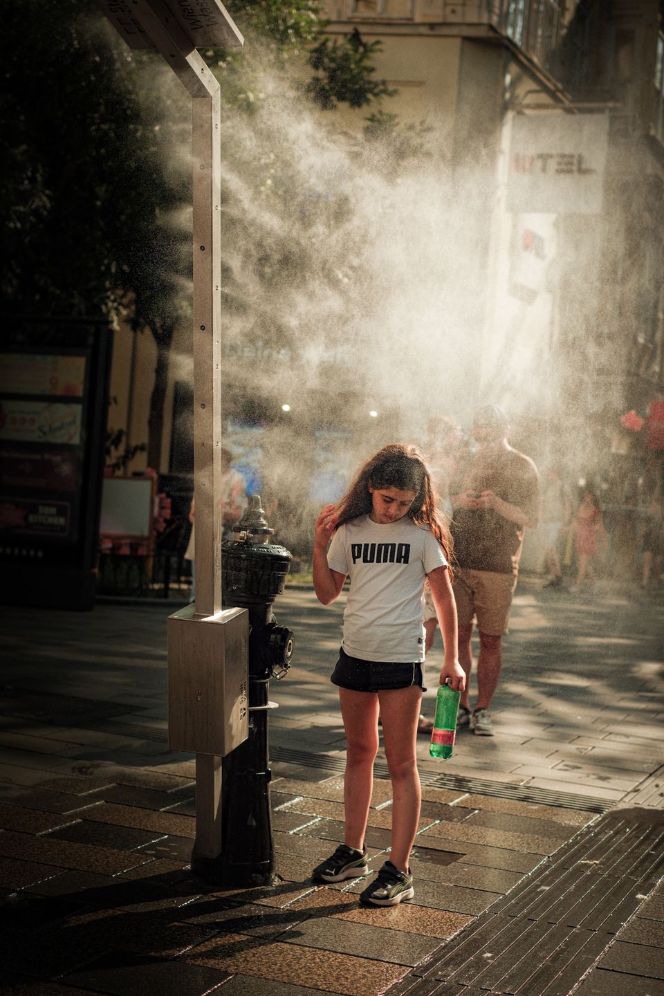 A girl stands under a misting fan on a sunny day in the city, cooling off