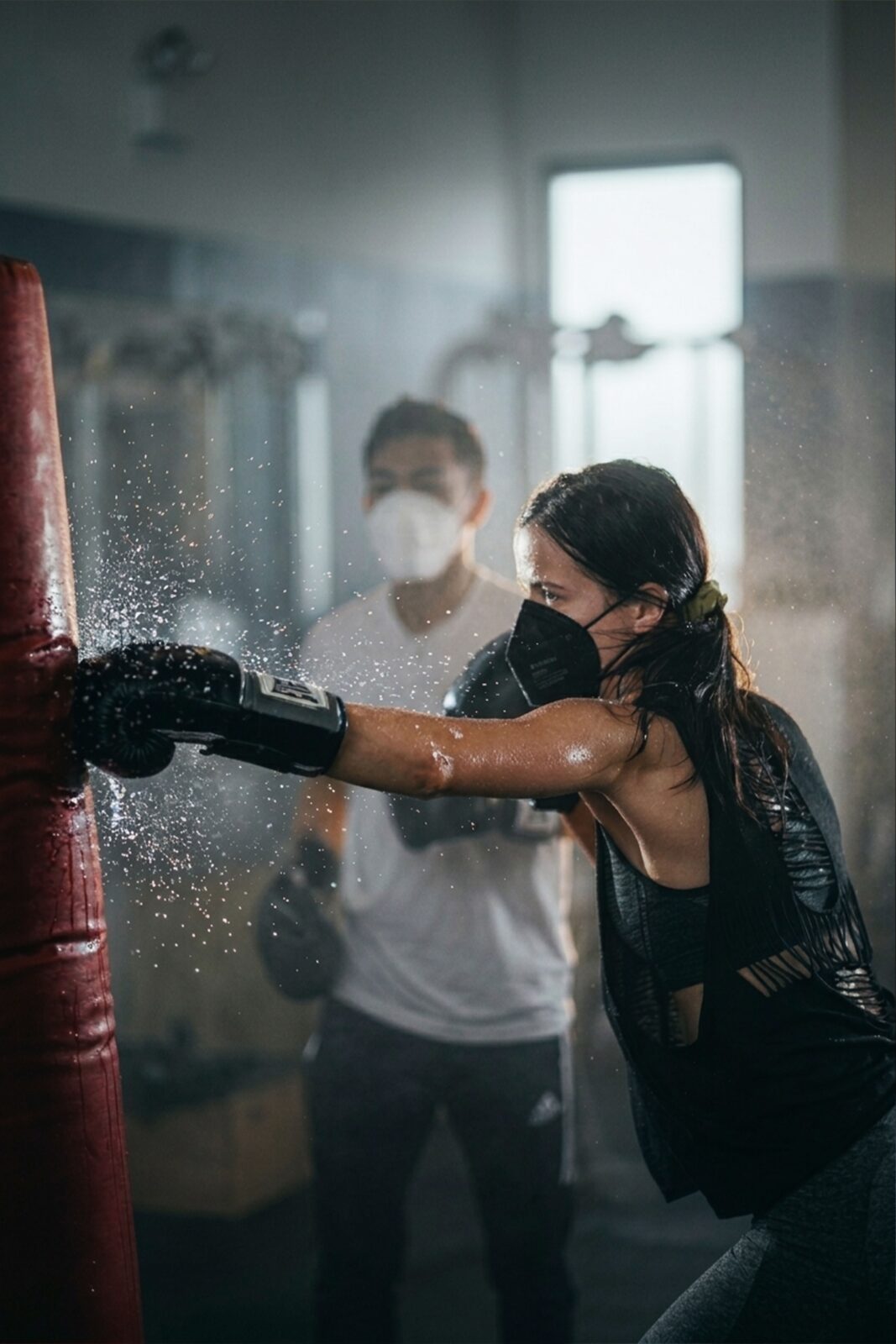A woman wearing a protective face mask and boxing gloves punching a heavy bag in a gym