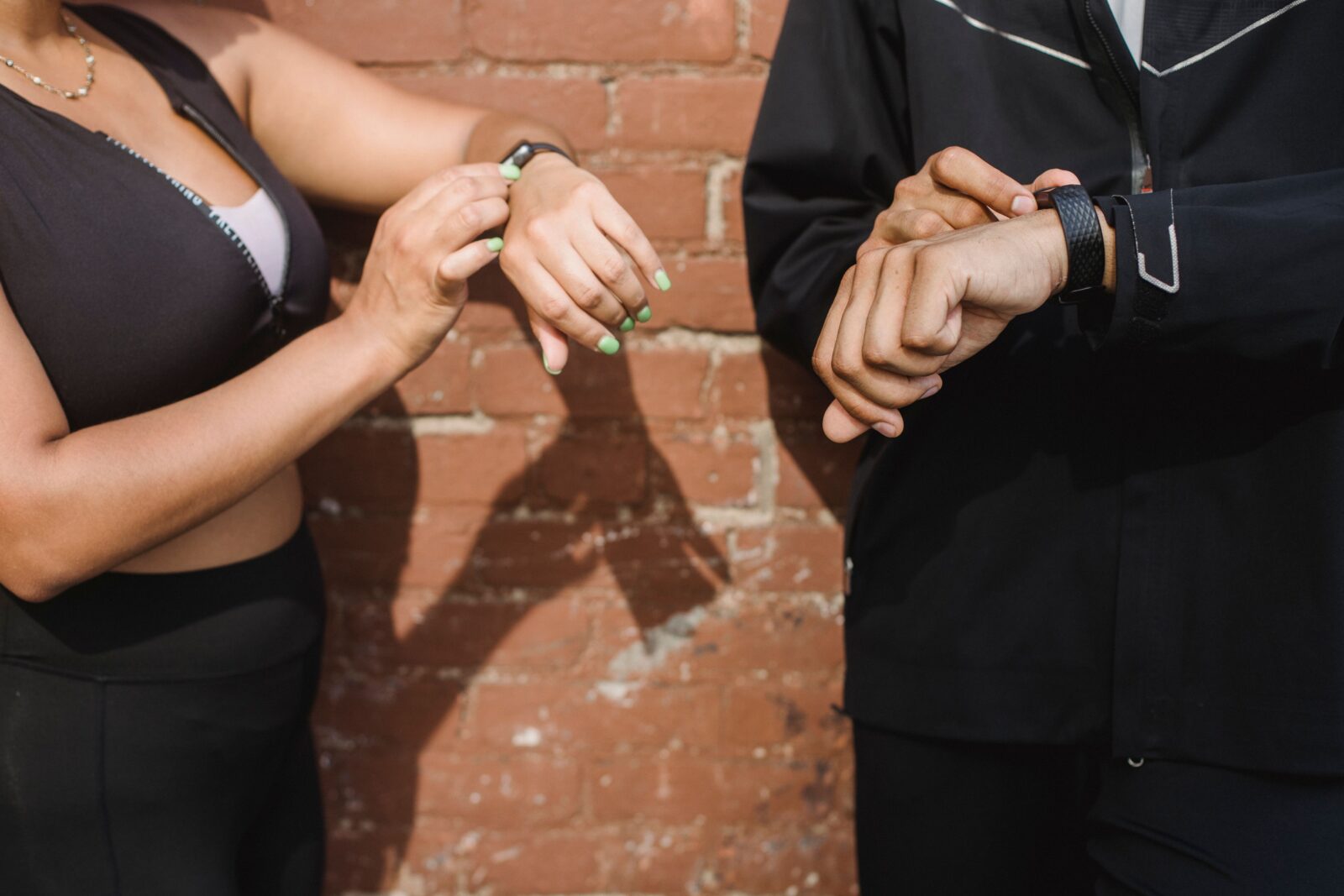 Two people in sportswear against a brick wall looking down at fitness trackers on their wrists