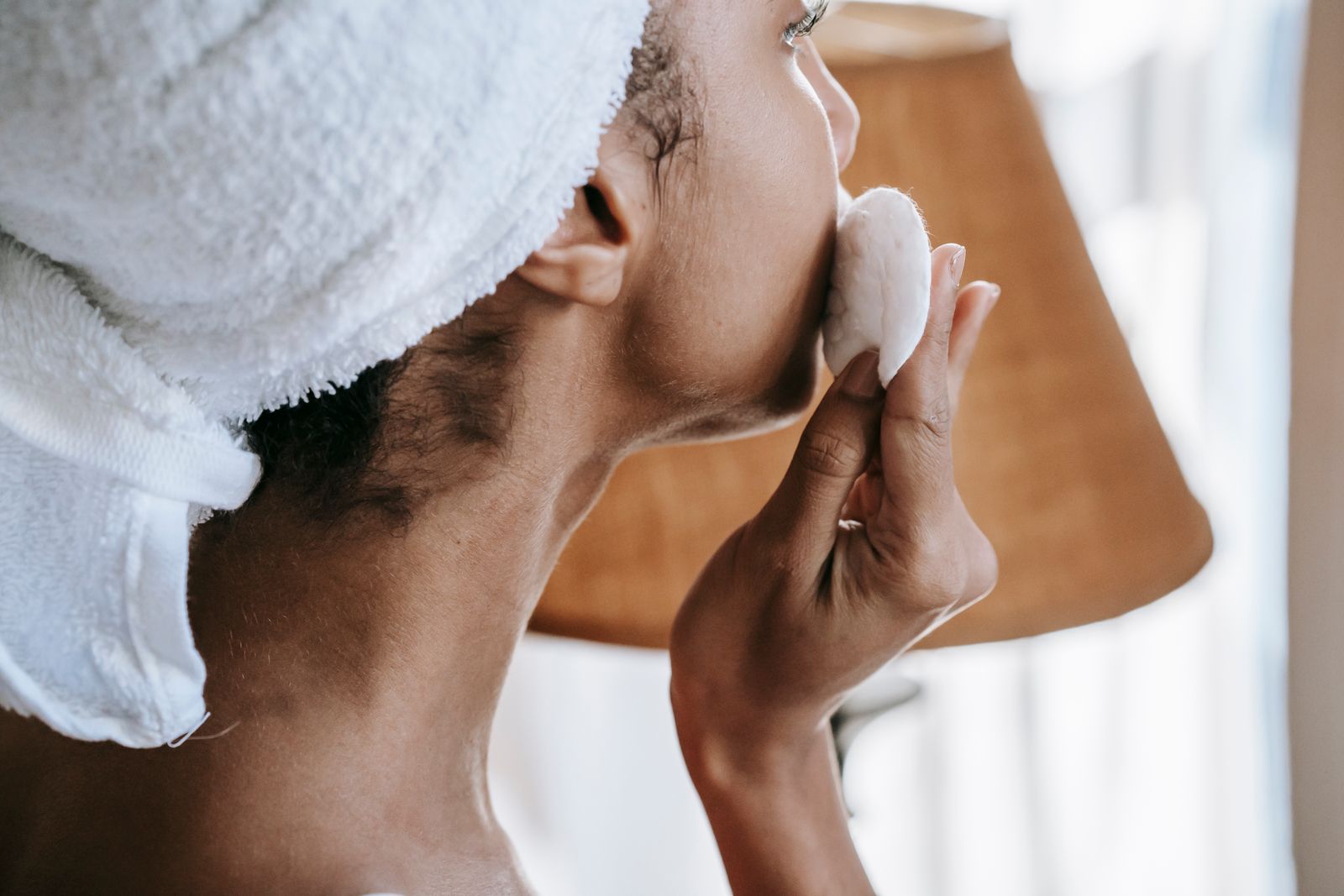 Woman with a towel wrapped around her head using a cotton round on her skin