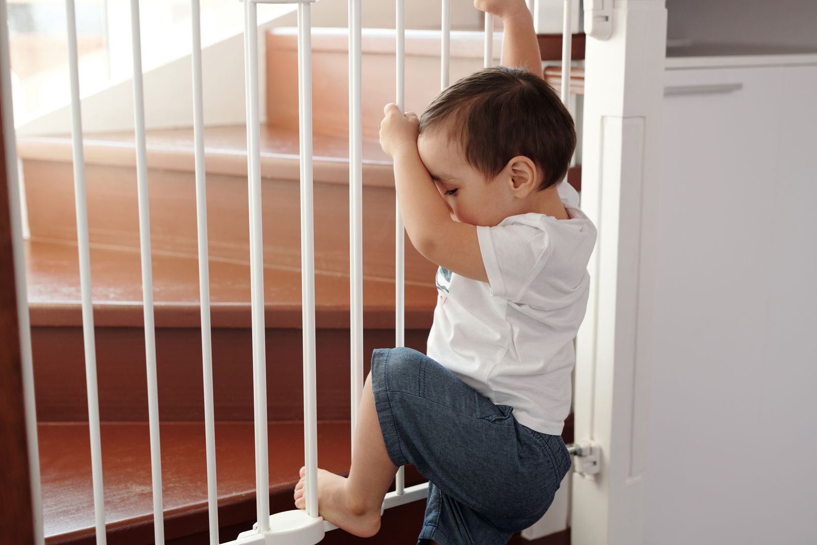 Toddler leaning against a white vertical-barred safety gate with a wooden staircase behind it