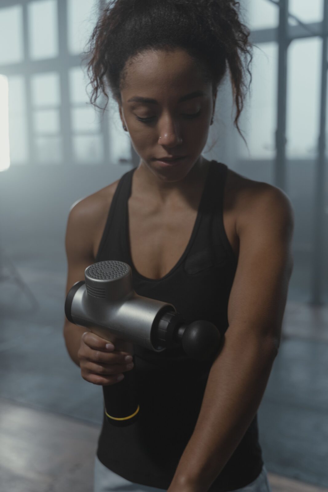A focused woman using a black massage gun on her arm in a gym