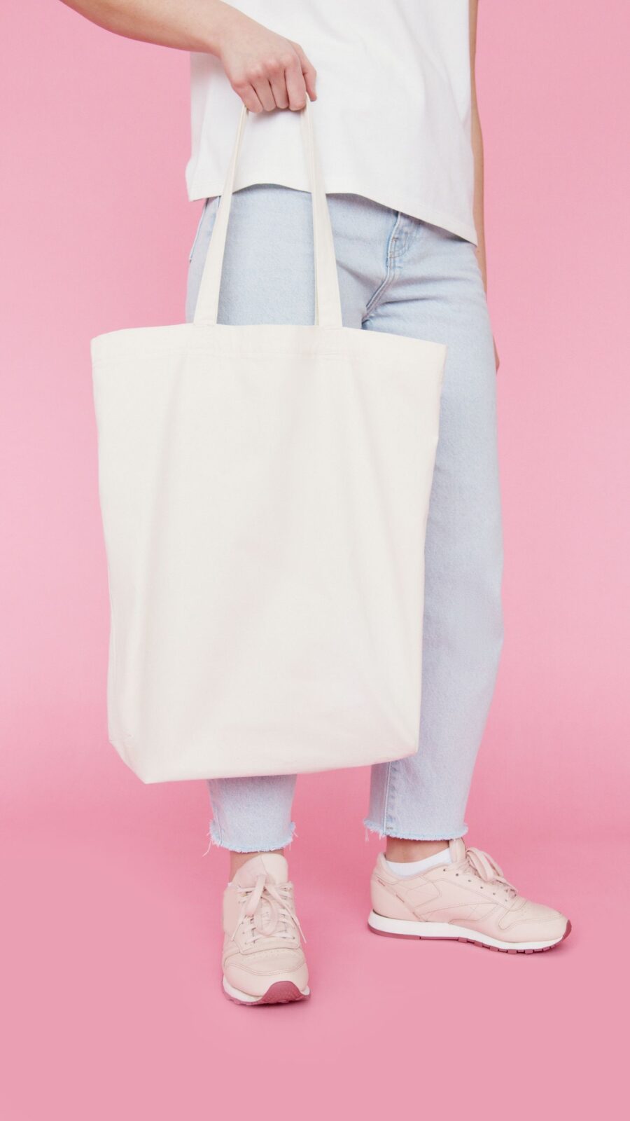 Person holding a plain unbranded cream canvas tote bag by both handles against a pink background