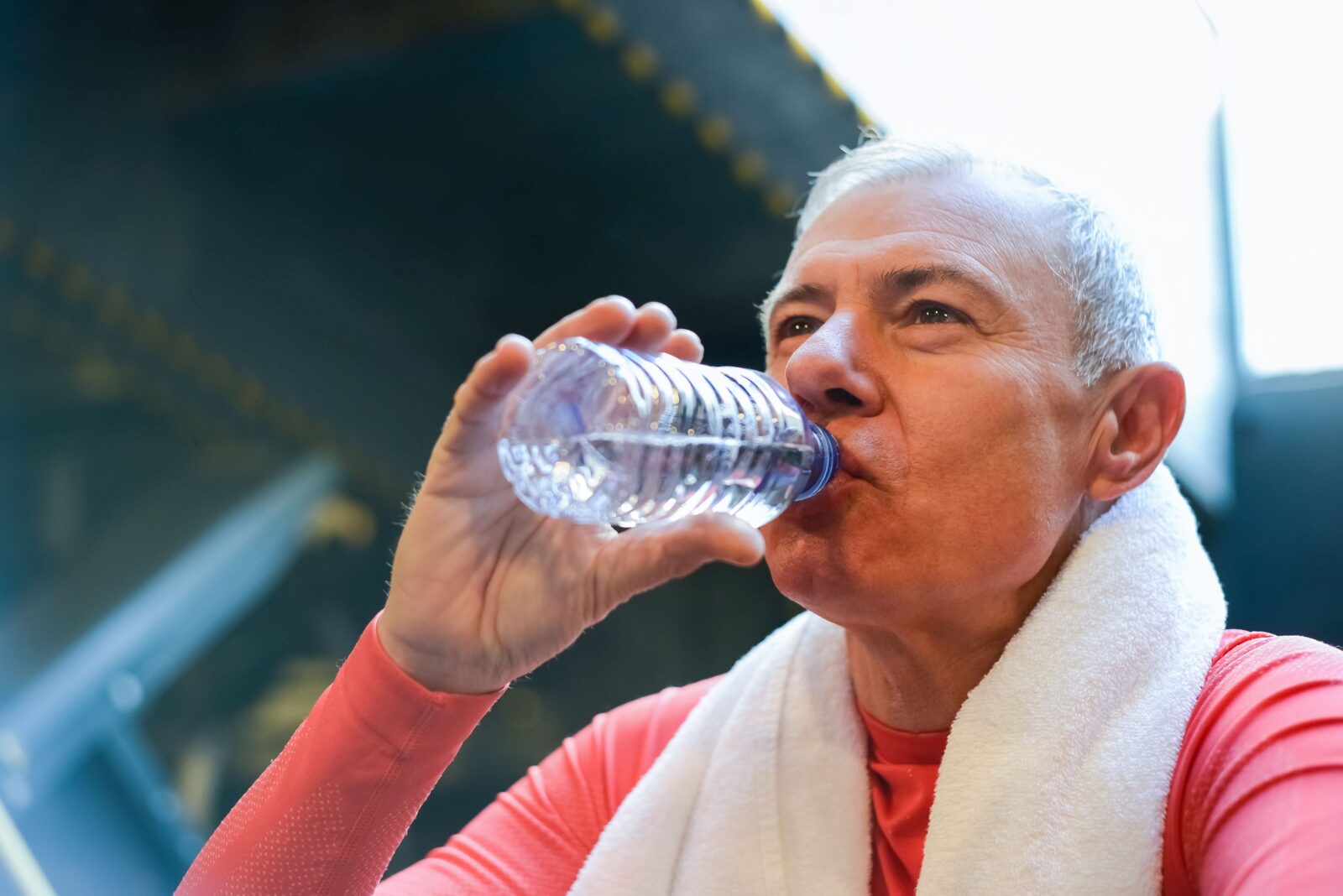 An elderly man with a towel around his neck drinking water after exercise