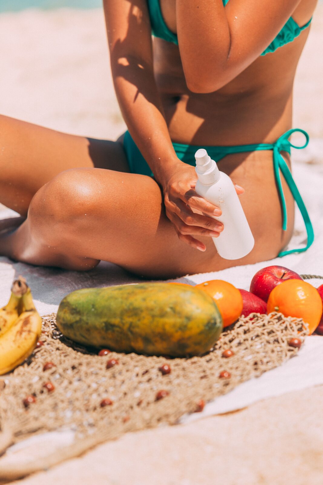 A woman sitting on a beach applying sunscreen from a spray bottle