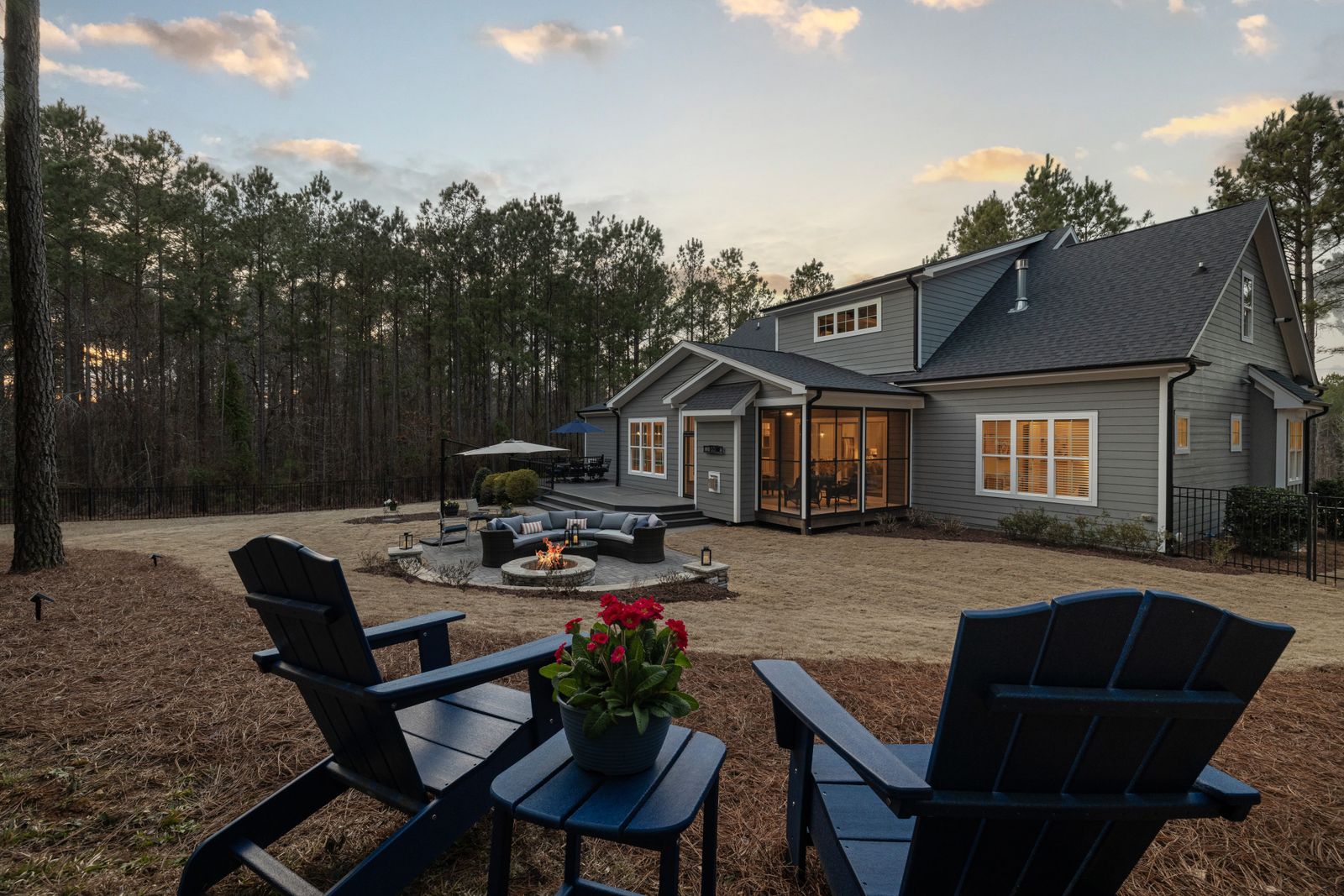 Adirondack chairs facing a stone fire pit seating area in a large backyard at dusk