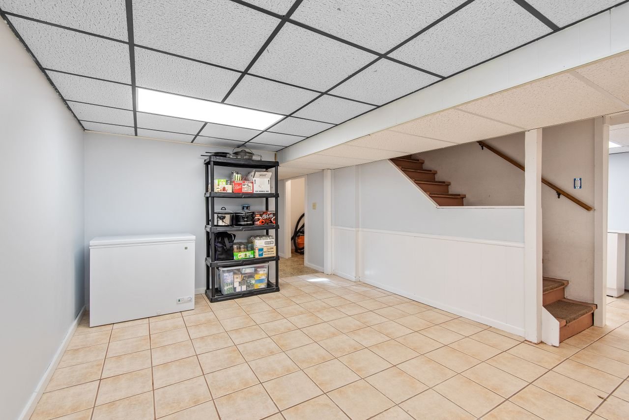 A finished basement with a tile floor, storage shelves, a chest freezer and a staircase