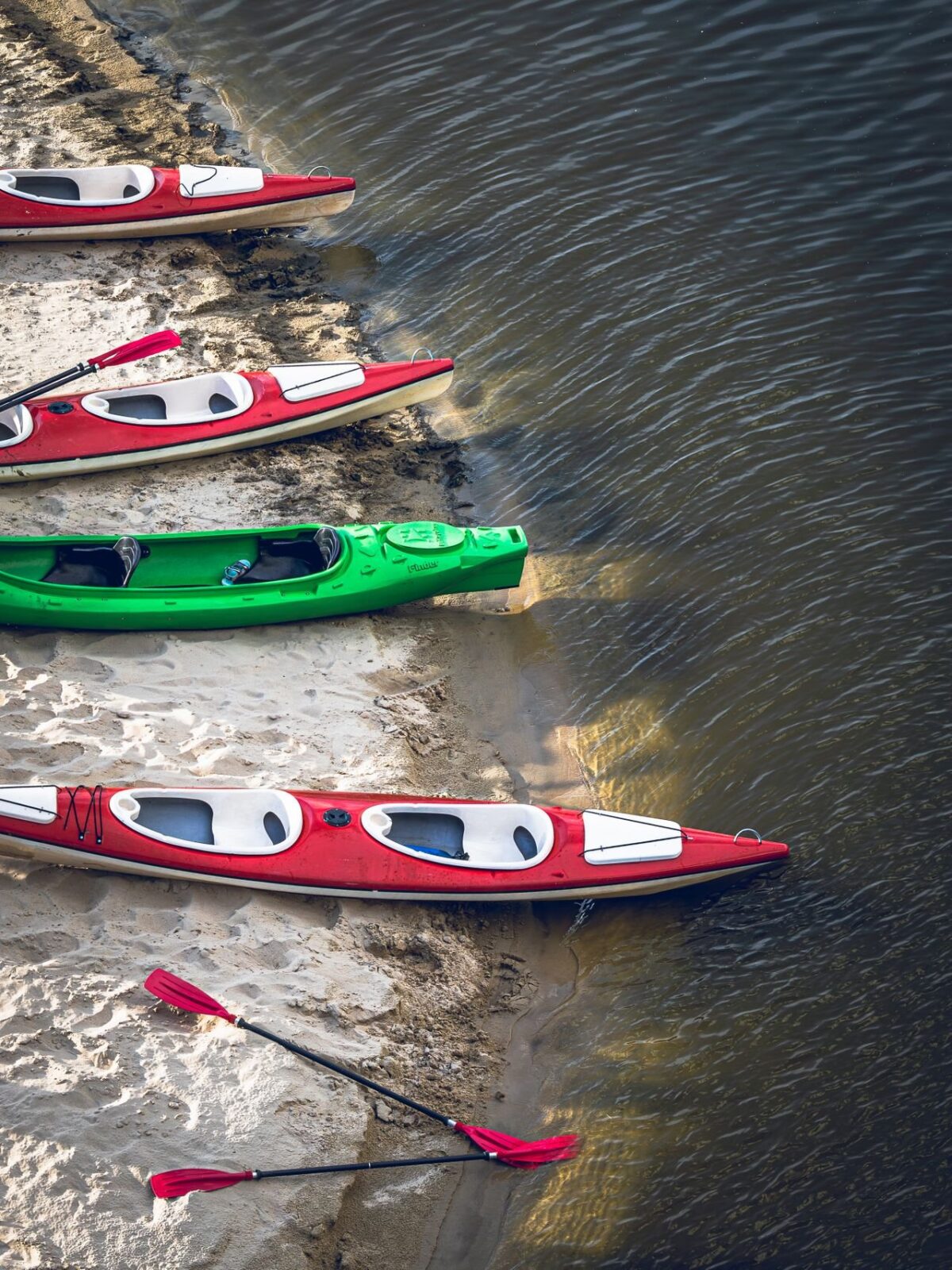 Red and green hardshell kayaks lined up on a sandy river shore, seen from above