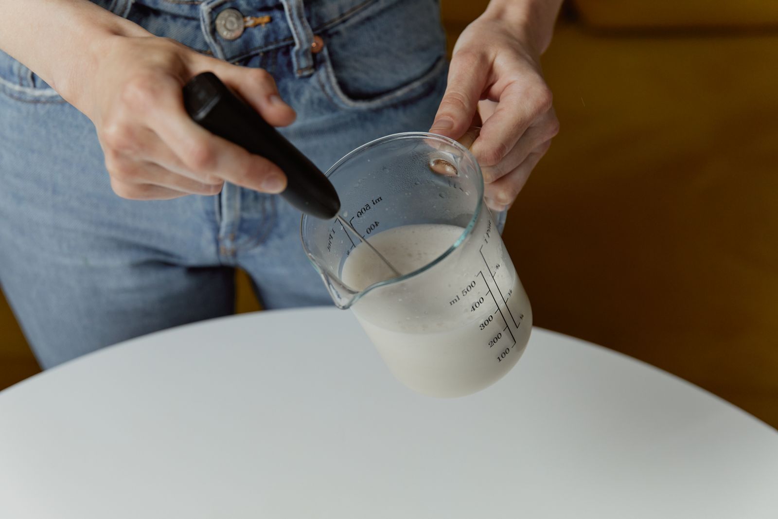 Hands frothing milk in a glass measuring cup with a battery-powered handheld whisk