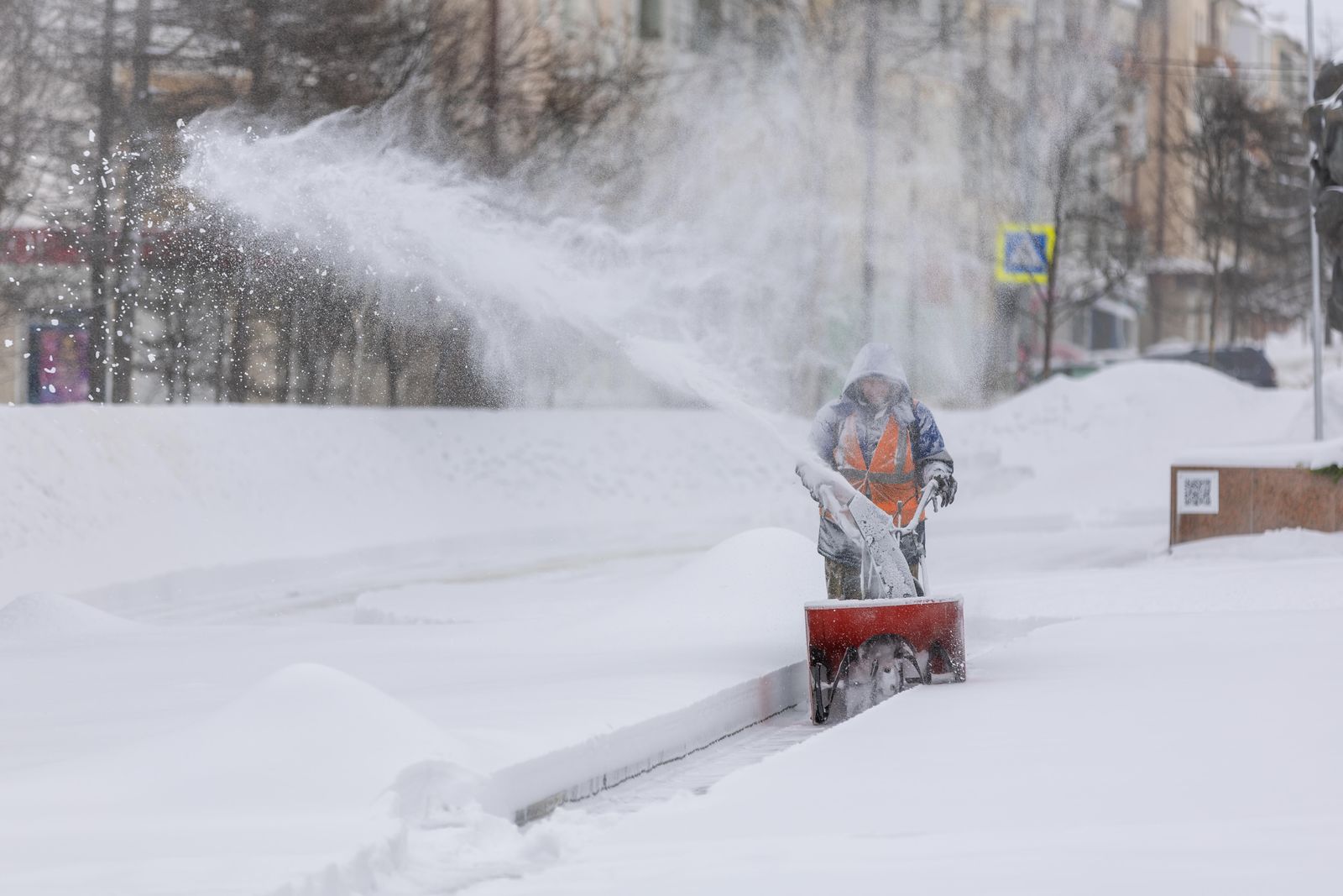 Hooded operator running a red walk-behind snow blower along a snow-covered street
