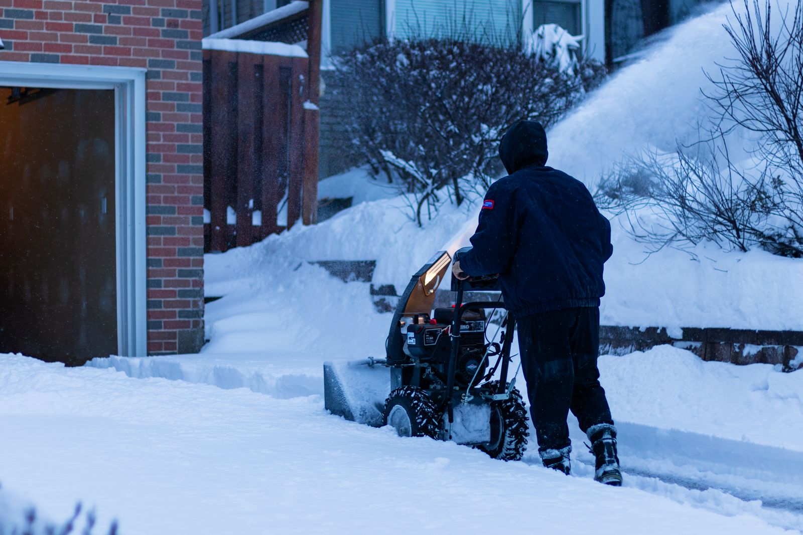 Person in a dark hooded jacket, seen from behind, clearing a snowy path with a snow blower beside a brick wall