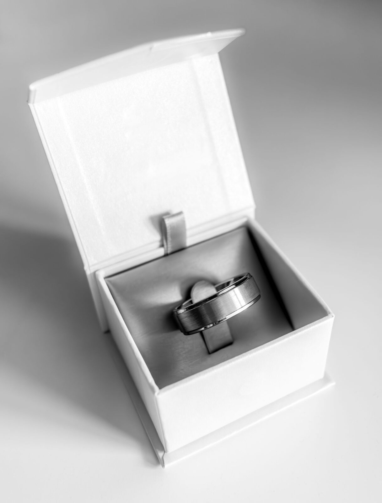 A single silver-colored band sitting in an open white ring box, in black and white