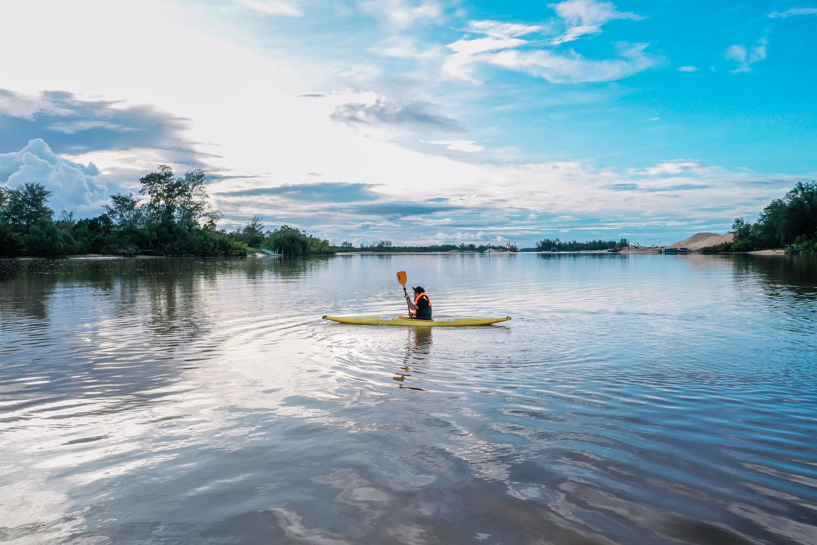 Distant kayaker in an orange life jacket paddling a yellow kayak on calm river water under a blue sky