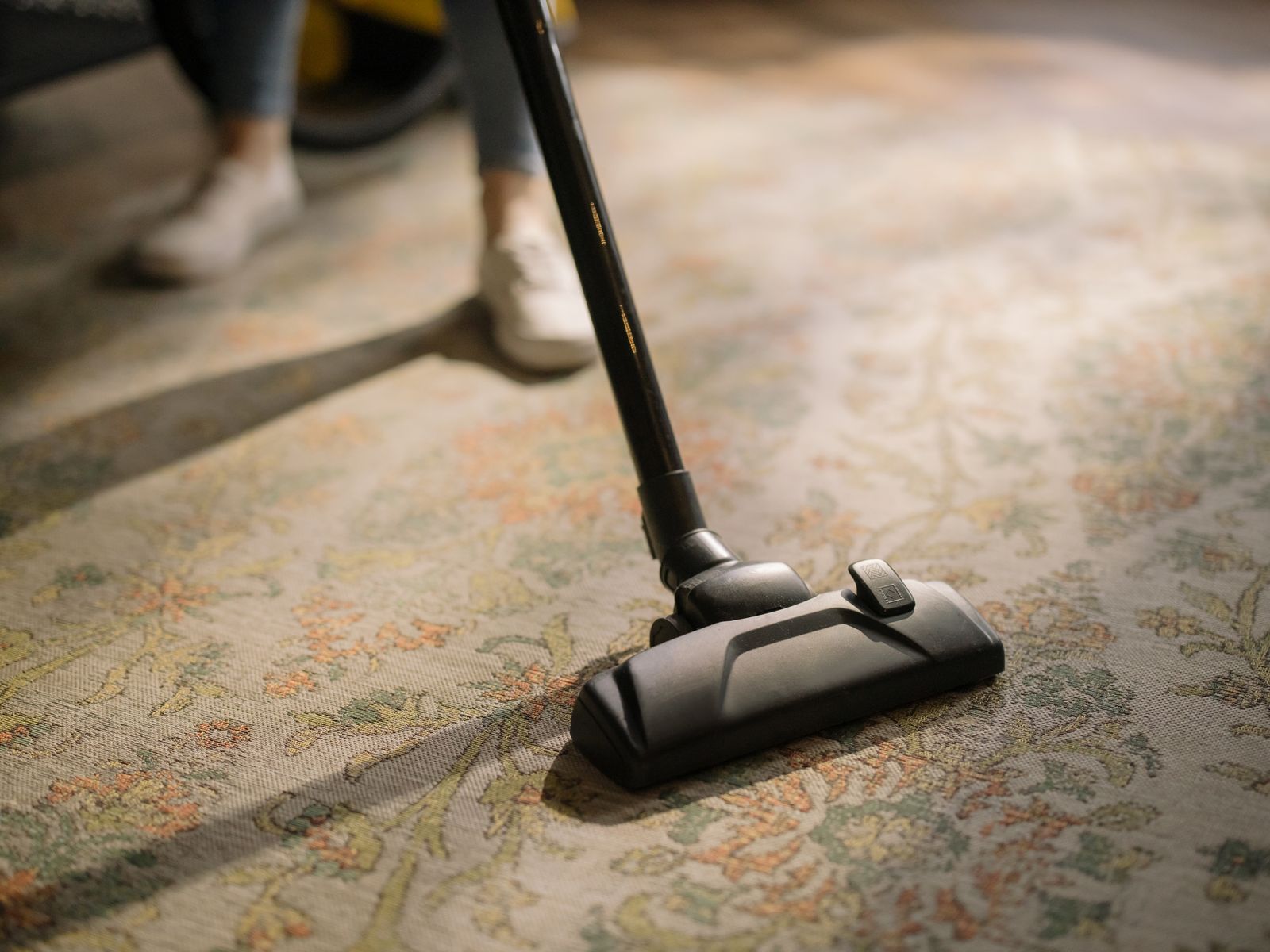 Black vacuum wand and floor head on a patterned area rug, a person’s feet behind it