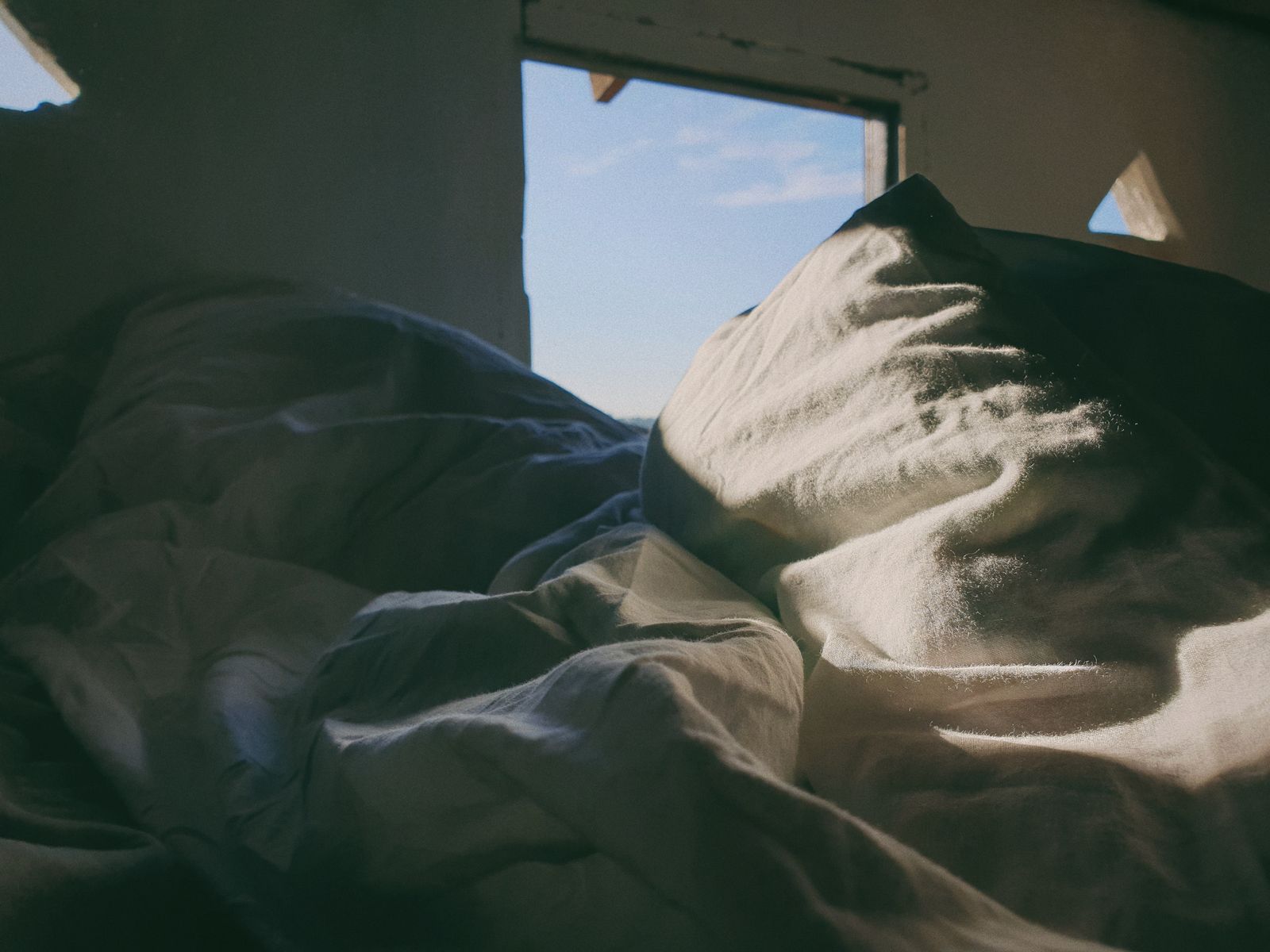 Rumpled white bedding and a pillow lit by daylight from a small window