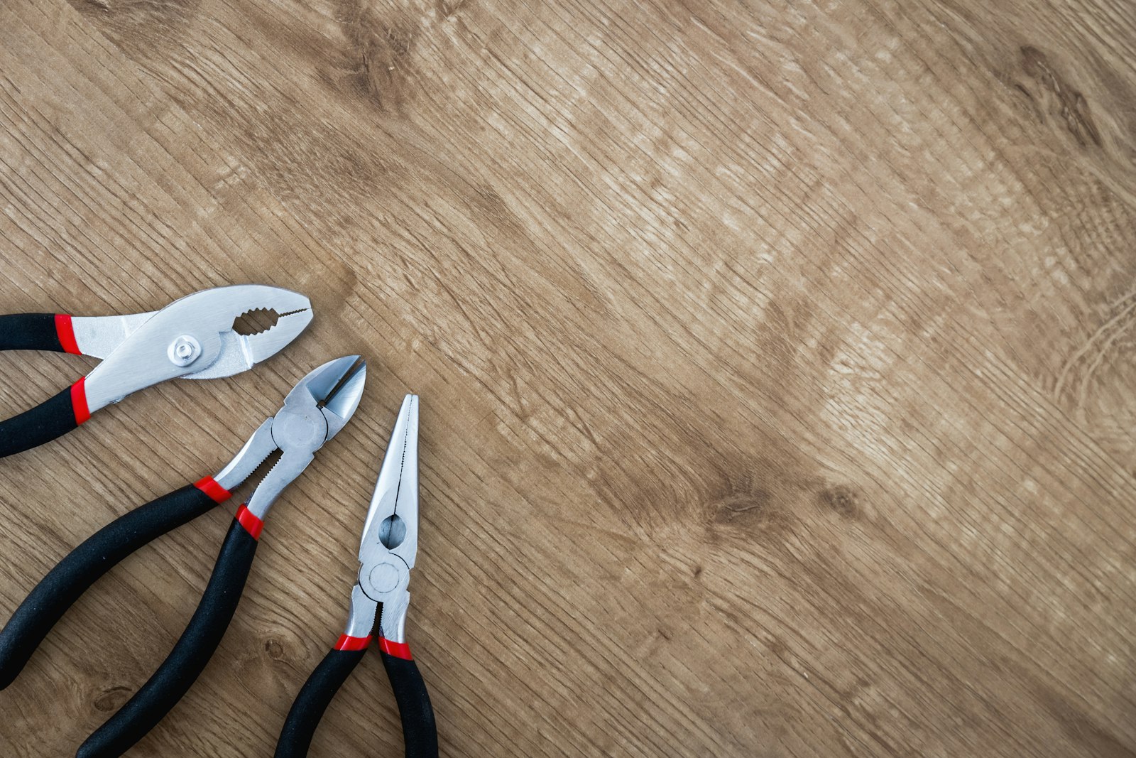 Slip-joint, diagonal cutting and needlenose pliers with black and red handles on a wooden surface