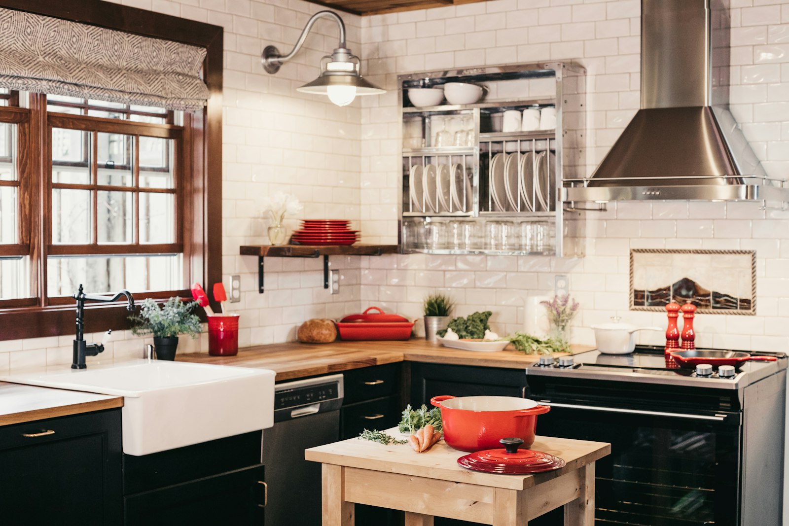 A dark freestanding range under a stainless hood in a white tiled kitchen with a red pot on the island