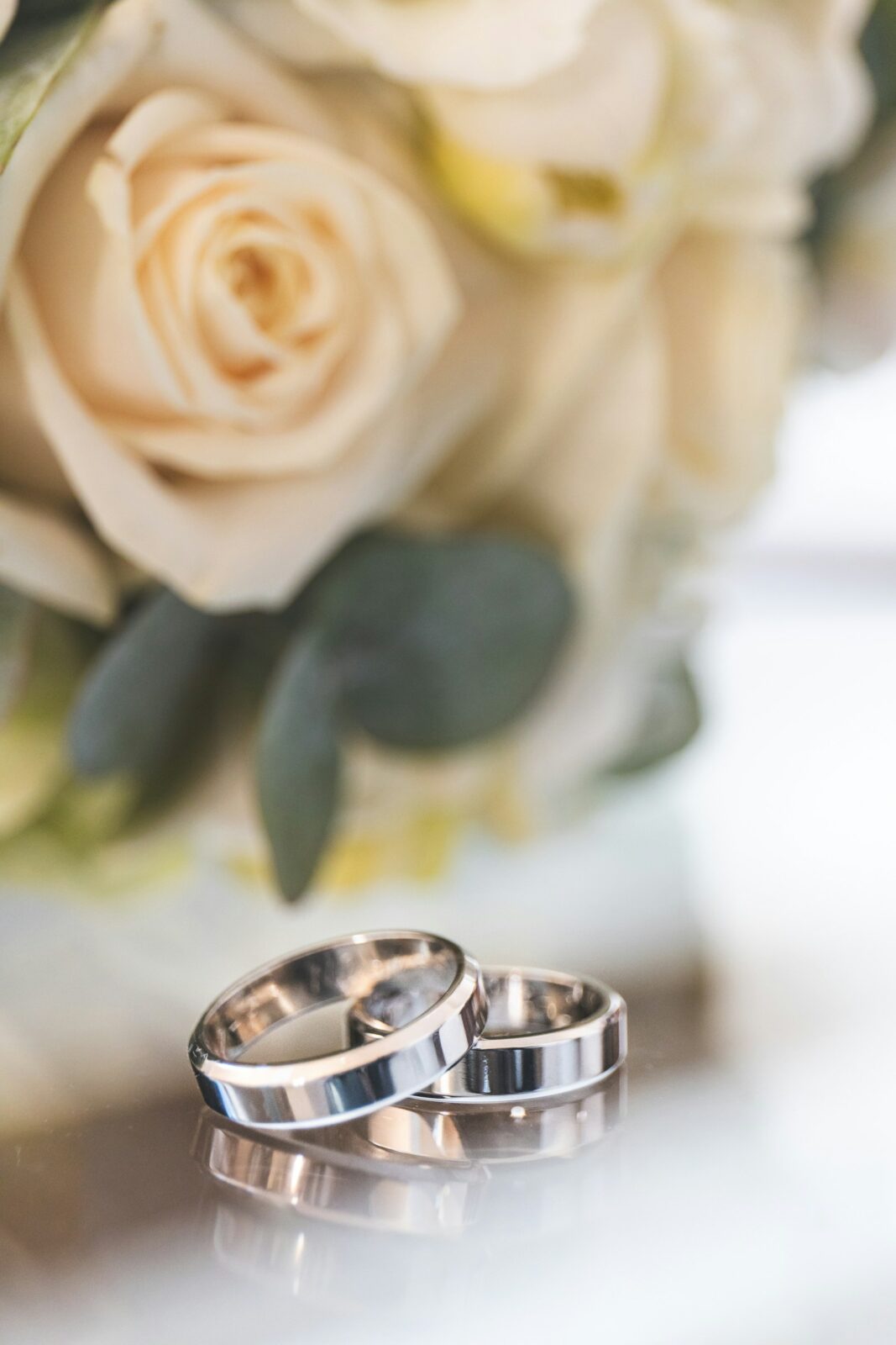 Two flat silver-colored wedding bands standing on a glossy surface in front of cream roses and green leaves