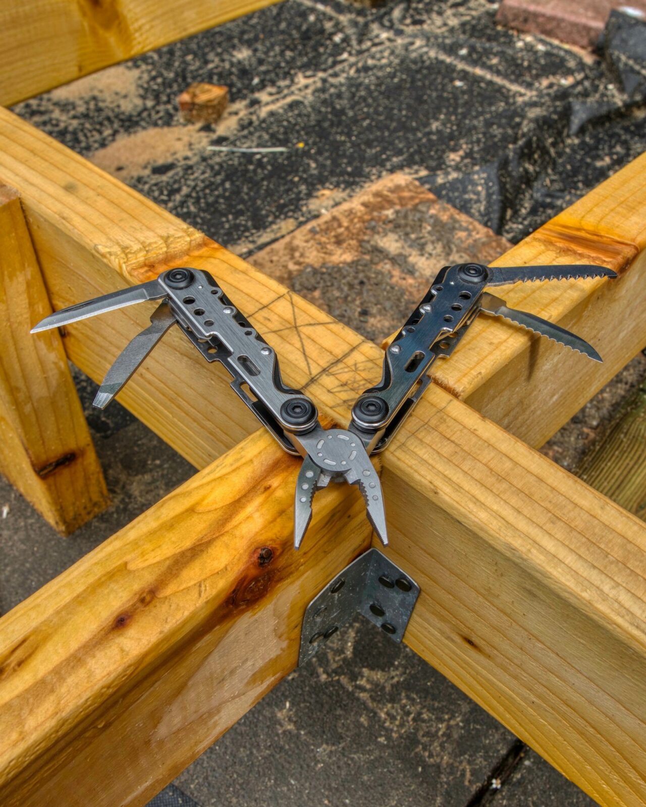 A gray multitool opened flat with pliers, blades and saw out, resting on the corner of a yellow lumber frame