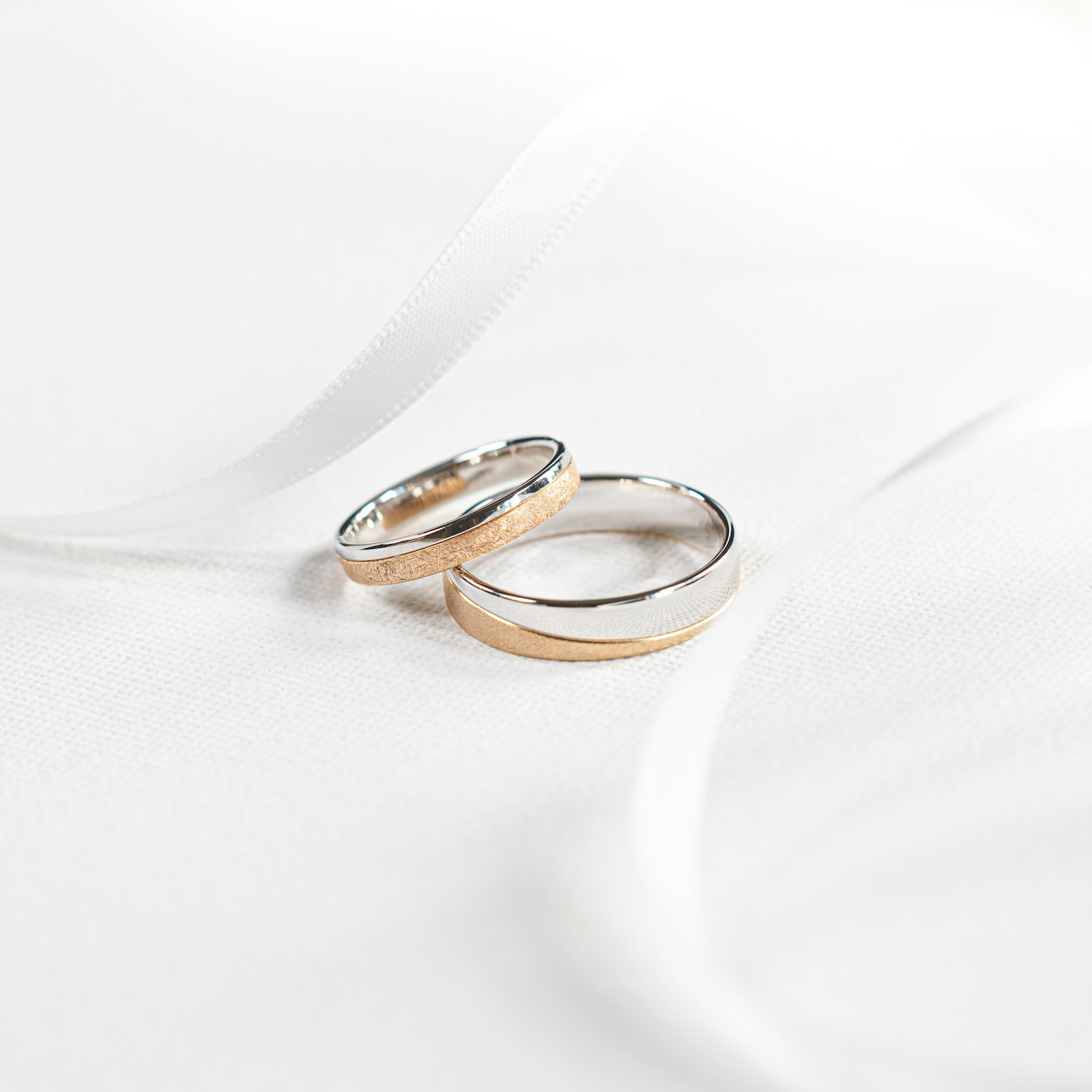 Two wedding bands, each half polished white metal and half textured rose-gold color, resting on white cloth