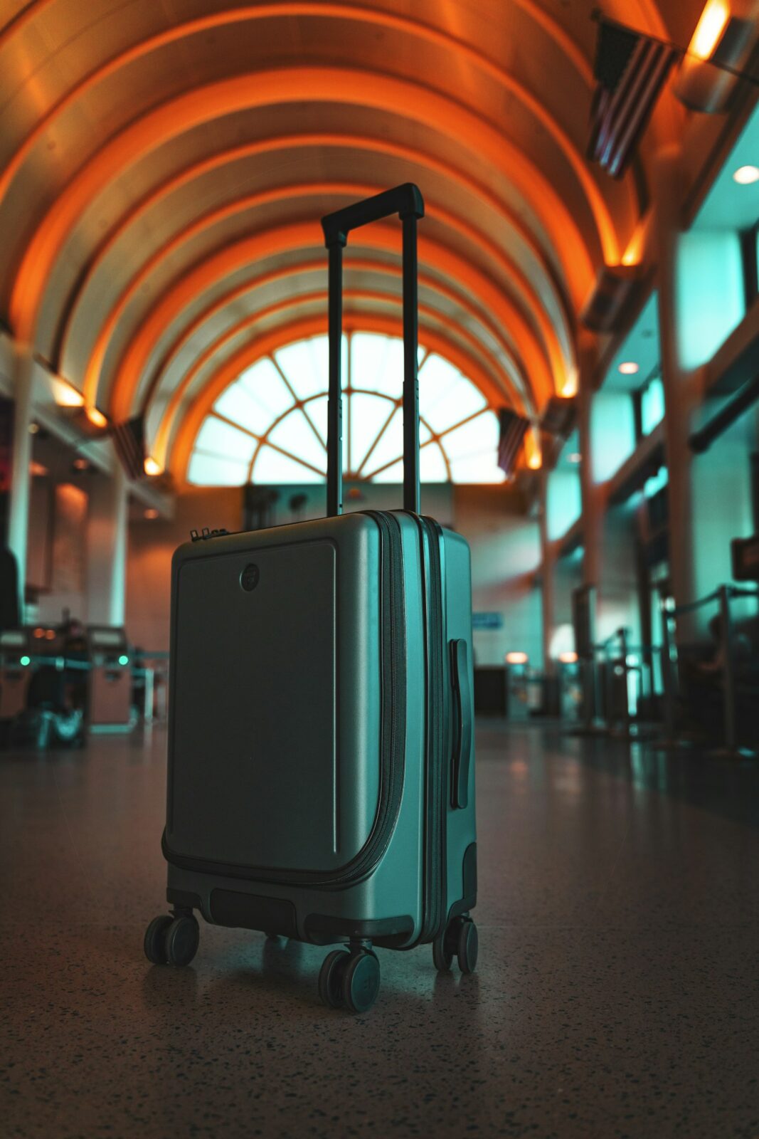 A green hardside carry-on suitcase with its handle raised on the floor of an arched airport terminal