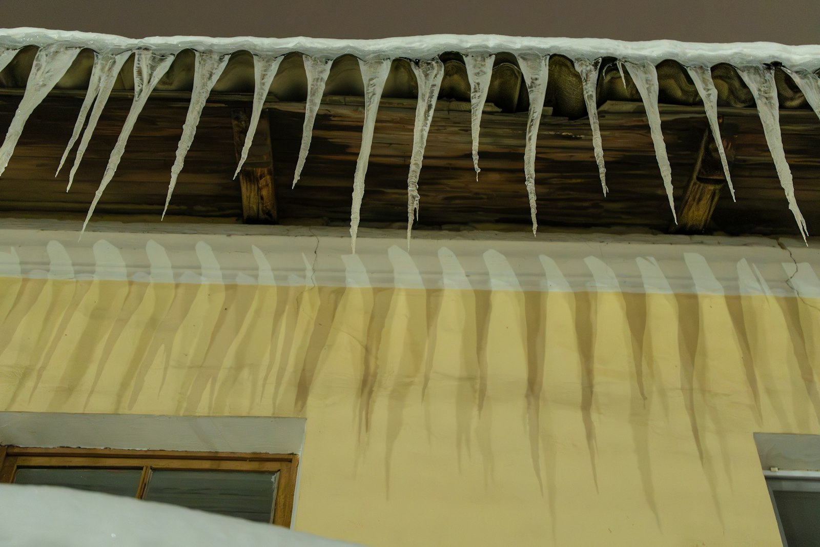 Long icicles under a snow-topped roof edge with dark water streaks down the wall below