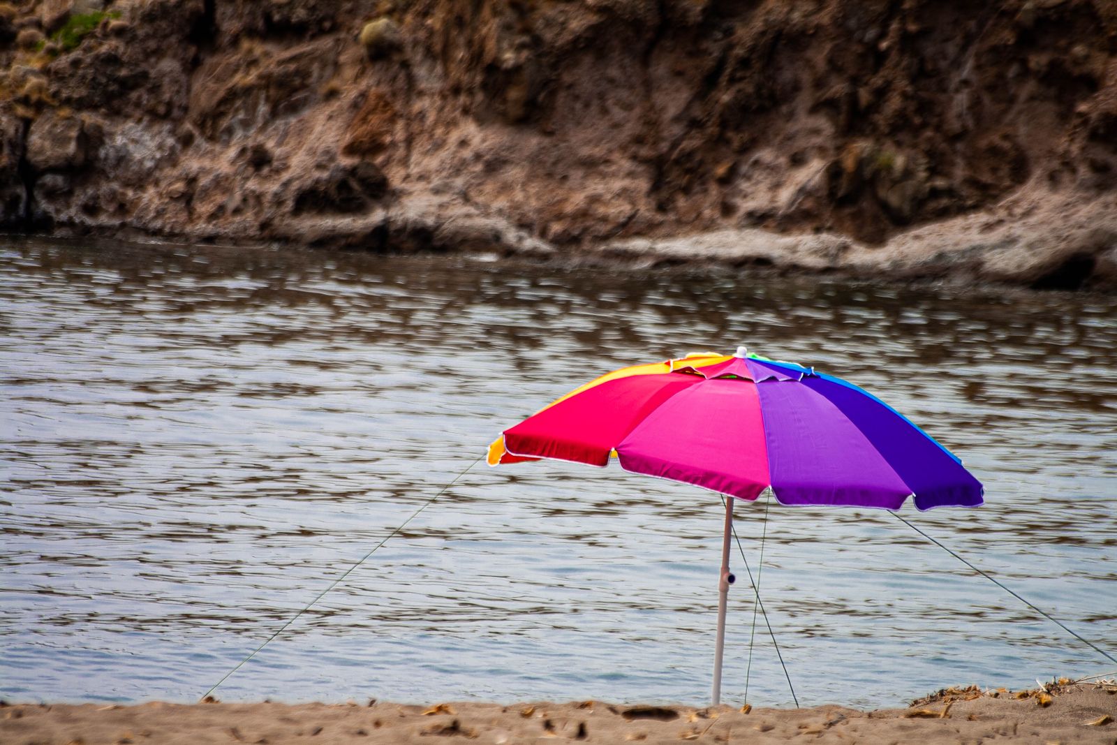 A rainbow-striped beach umbrella planted in the sand at the water's edge