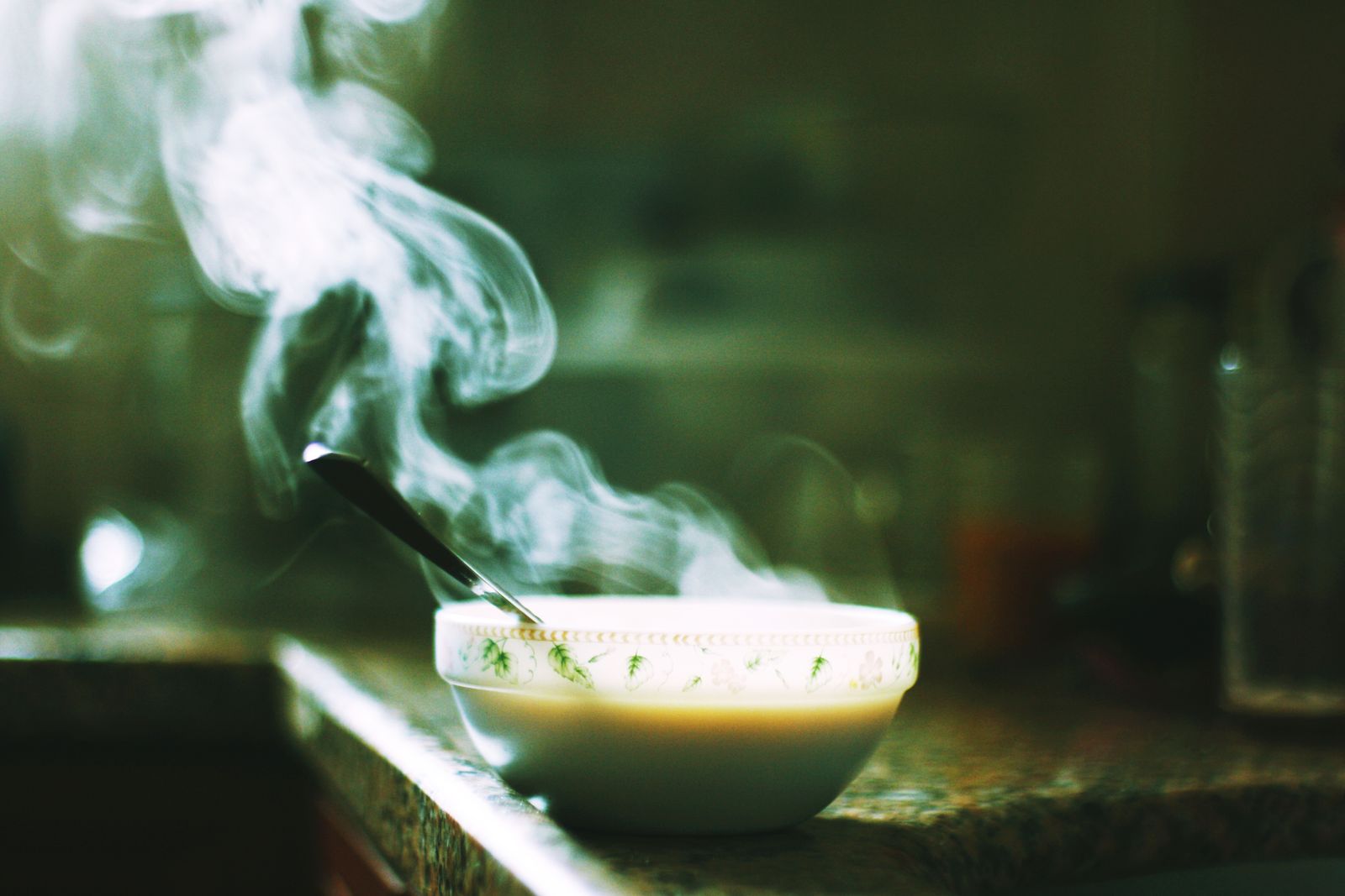 A white bowl of hot soup with a spoon, steam rising, on a dark kitchen counter